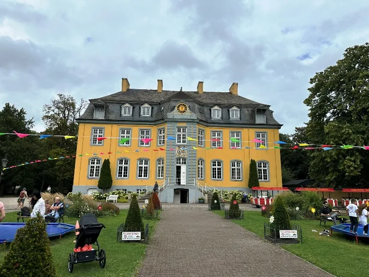 Das Bild zeigt Schloss Beck bei Dorsten, eine barocke Schlossanlage umgeben von einer gepflegten Gartenlandschaft. Das Hauptgebäude aus hellem Stein wird durch symmetrische Flügelbauten ergänzt. Im Vordergrund ist ein weitläufiger Rasen zu sehen, während dahinter eine Baumreihe und ein klarer Himmel das Schloss einrahmen.